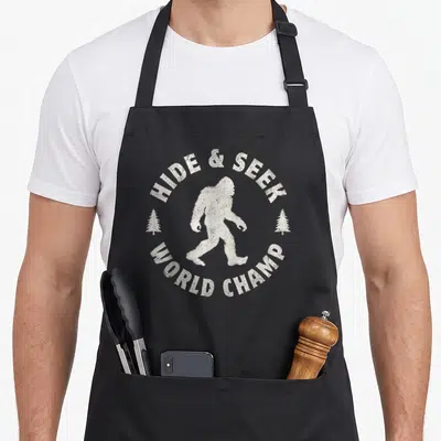 Hide seek bigfoot apron - White distressed Bigfoot walking between two trees with arched text HIDE & SEEK WORLD CHAMP — shown on a black cotton canvas apron against a white background