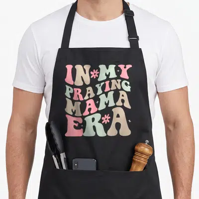Praying mother's day aprons - IN MY PRAYING MAMA ERA in wavy retro font with pink flowers and beige stars on black — shown on a black cotton canvas apron against a white background