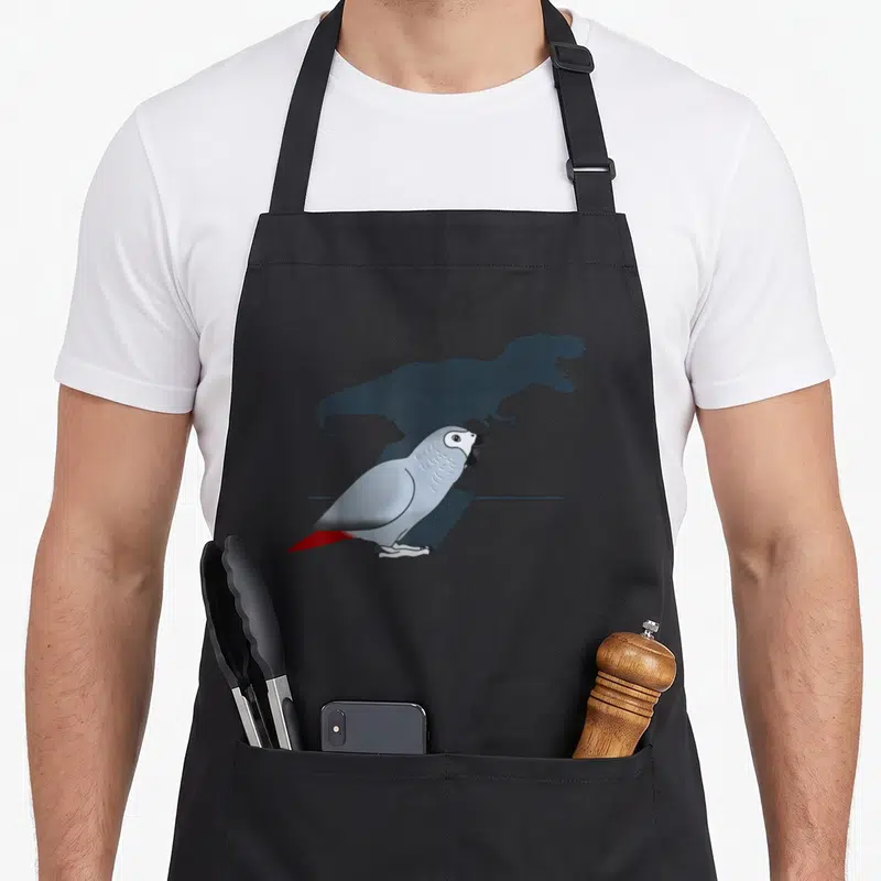 Birb african aprons - Grey parrot with red tail and open beak casting a dinosaur shadow on grey wall — shown on a black cotton canvas apron against a white background