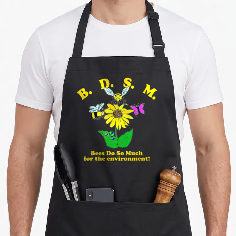 Bee b and m apron - Sunflower with bees, butterfly, caterpillar and B.D.S.M. text in yellow, green, purple — shown on a black cotton canvas apron against a white background