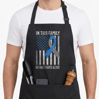 Flag family aprons - Blue ribbon over distressed US flag with text IN THIS FAMILY NO ONE FIGHTS ALONE — shown on a black cotton canvas apron against a white background