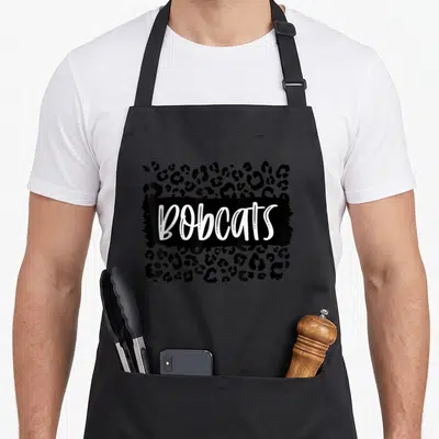 Bobcat leopard print apron - White BOBCATS script on black brushstroke over black leopard print pattern — shown on a black cotton canvas apron against a white background