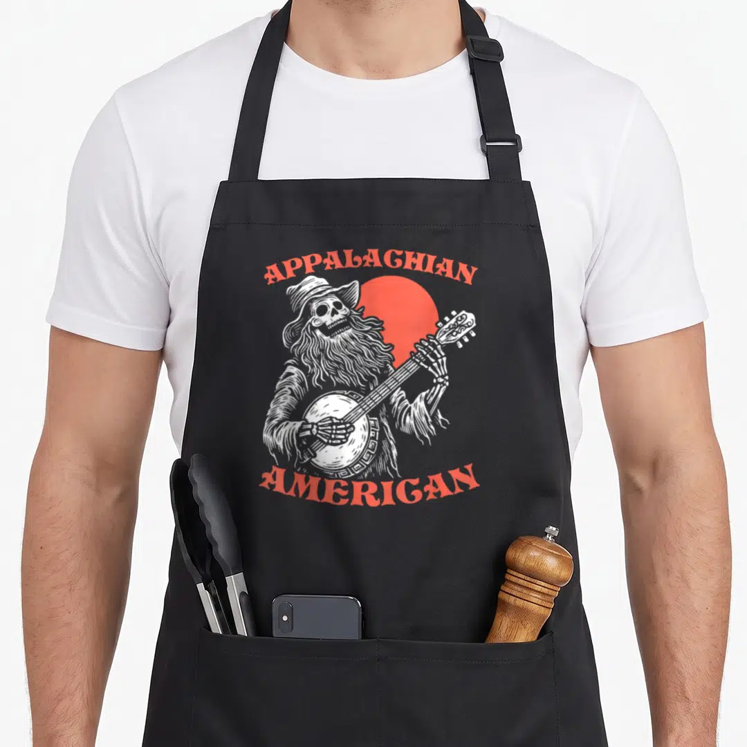 Appalachian skeleton apron - White line-art skeleton playing banjo in front of red sun with APPALAGHIAN AMERICAN text — shown on a black cotton canvas apron against a white background