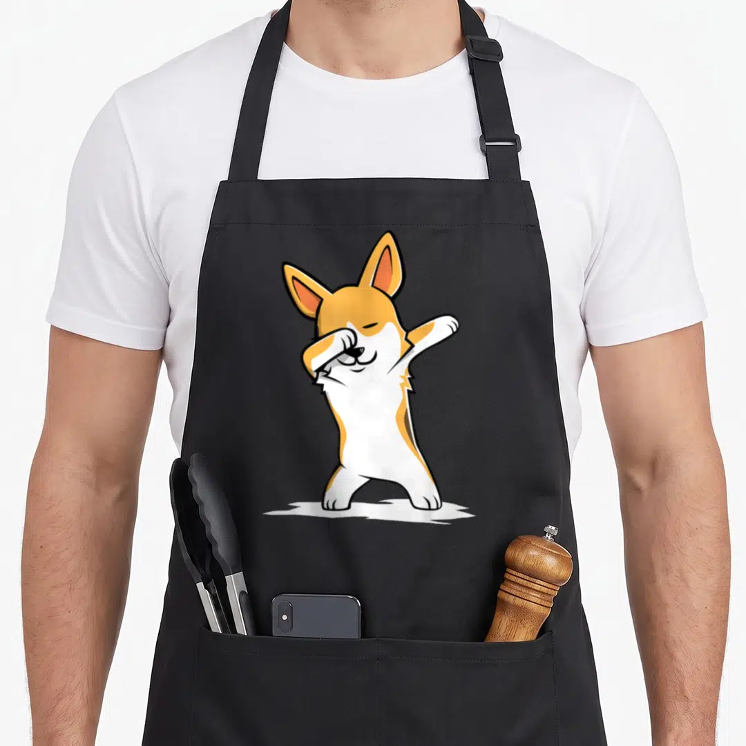 Dabbing corgi apron - Cartoon corgi in orange and white standing and dabbing with one paw over its eye — shown on a black cotton canvas apron against a white background
