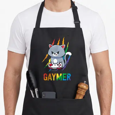 Gaymer video game apron - Grumpy grey cat on white controller with rainbow streaks and GAYMER text below — shown on a black cotton canvas apron against a white background