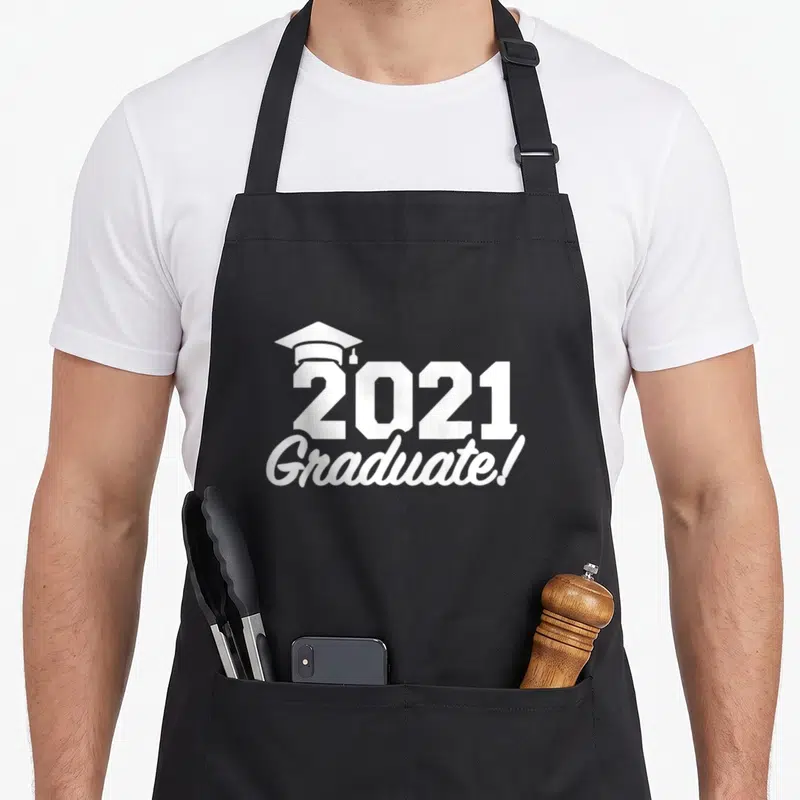 Funny graduate apron - White 2021 graduation cap and Graduate text typography design — shown on a black cotton canvas apron against a white background
