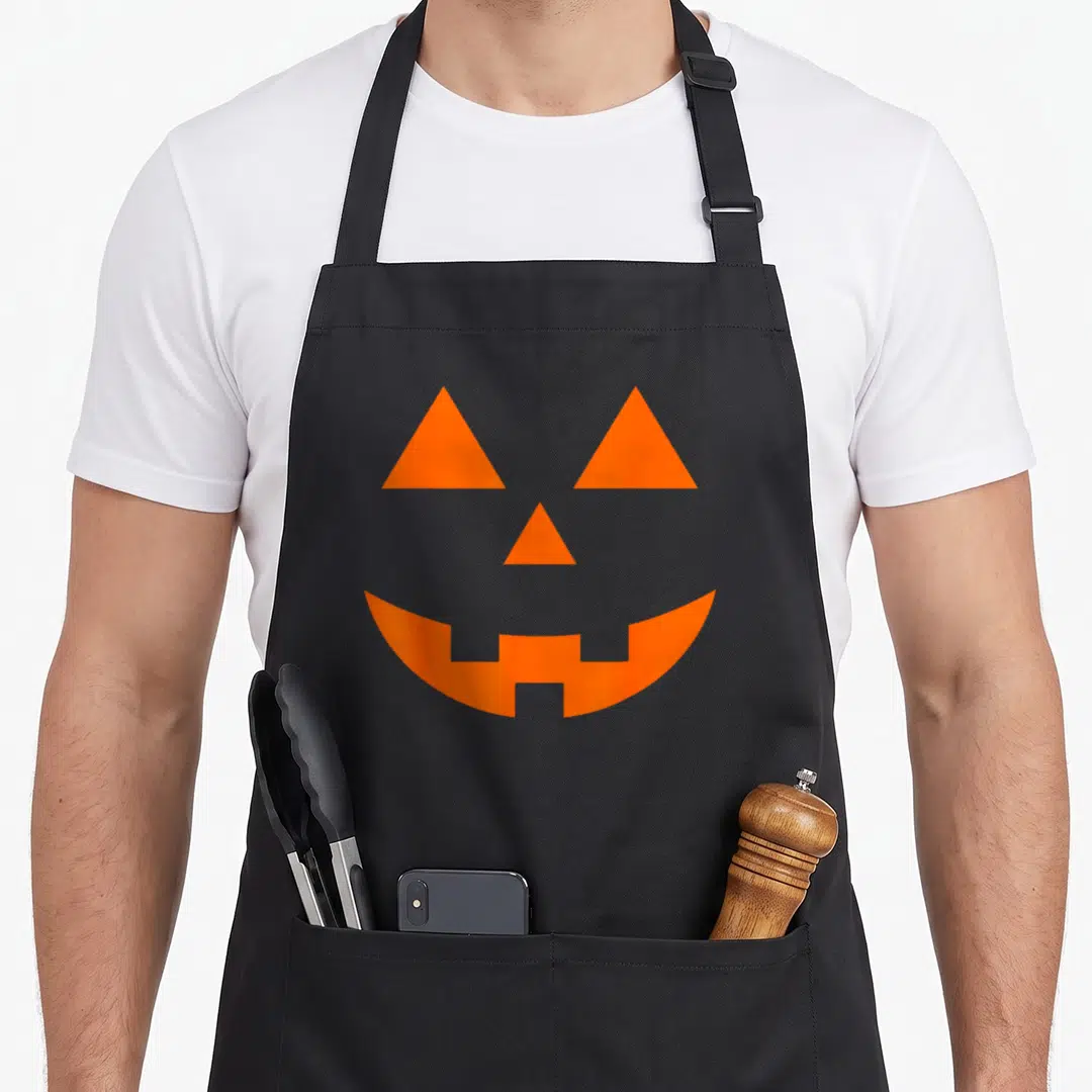 Jack spooky apron - Orange minimalist jack o lantern face with triangular eyes and toothy crescent mouth on black — shown on a black cotton canvas apron against a white background