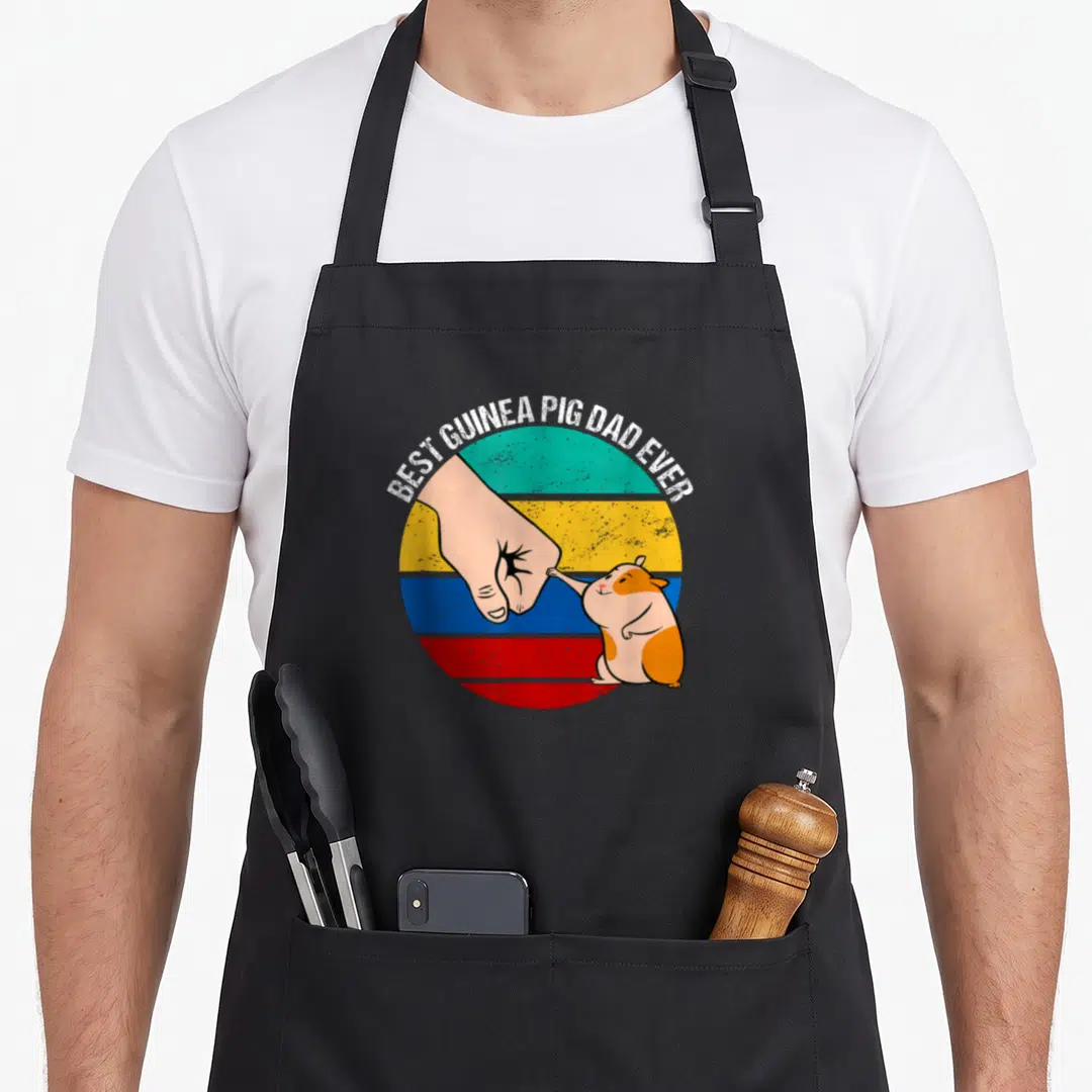 Pig guinea dad apron - Cartoon hand and guinea pig fist bumping inside a teal yellow blue and red striped circle — shown on a black cotton canvas apron against a white background