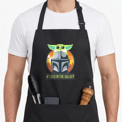 Galaxy fathers day apron - Grogu peeking over Mandalorian helmet with #1 DAD IN THE GALAXY text — shown on a black cotton canvas apron against a white background