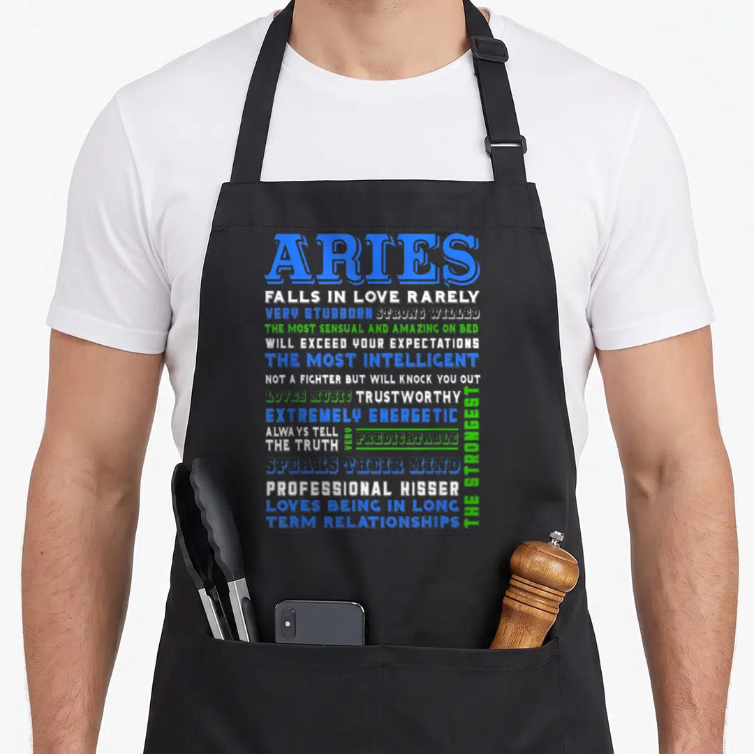Characteristic aries kitchen apron - Stacked blue white and green text listing Aries zodiac traits like stubborn — shown on a black cotton canvas apron against a white background
