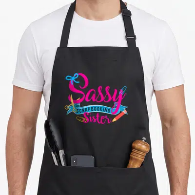 Sassy crafting apron - Sassy Scrapbooking Sister text with scissors, pencil, paperclips, and blue ribbon in pink — shown on a black cotton canvas apron against a white background