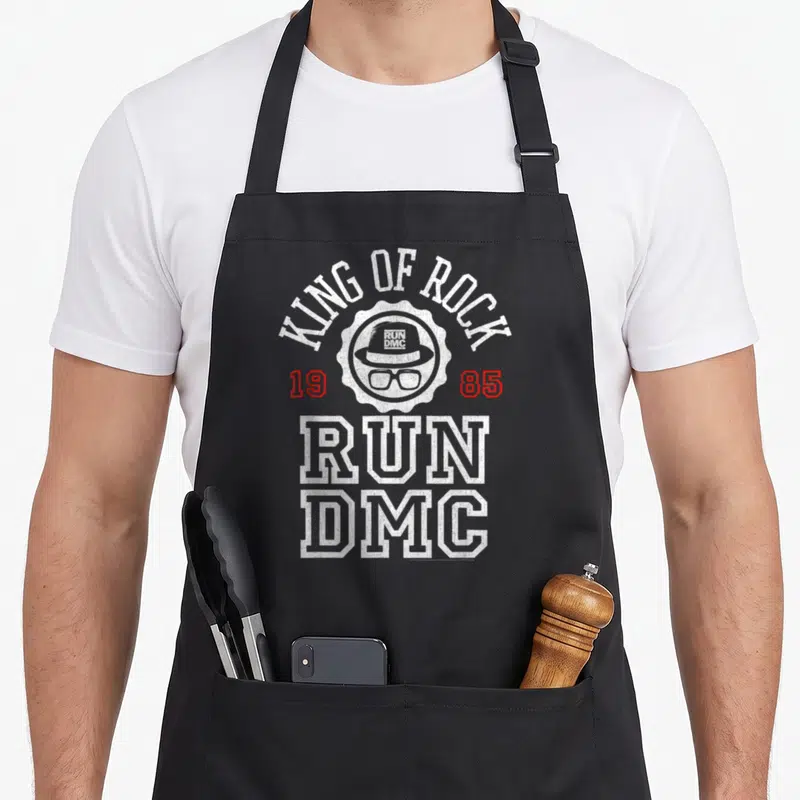 Dmc king apron - KING OF ROCK text arching over a hat and glasses seal with 1985 dates — shown on a black cotton canvas apron against a white background