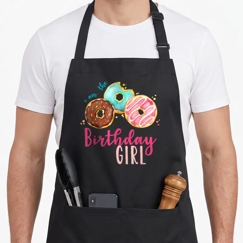 Theme family cooking aprons - Three watercolor donuts in pink blue brown yellow with i am the Birthday GIRL text — shown on a black cotton canvas apron against a white background