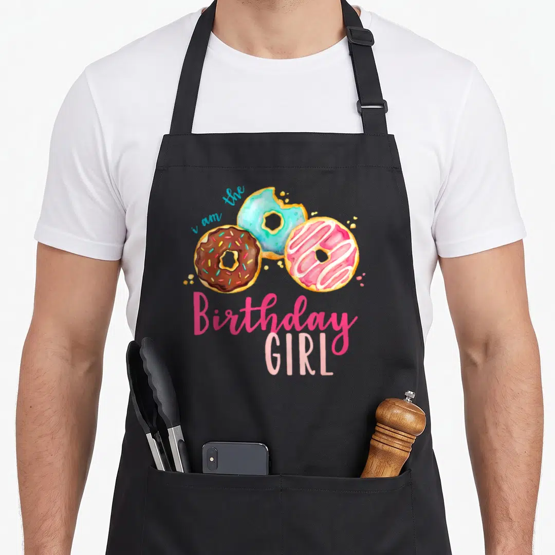 Theme family cooking aprons - Three watercolor donuts in pink blue brown yellow with i am the Birthday GIRL text — shown on a black cotton canvas apron against a white background