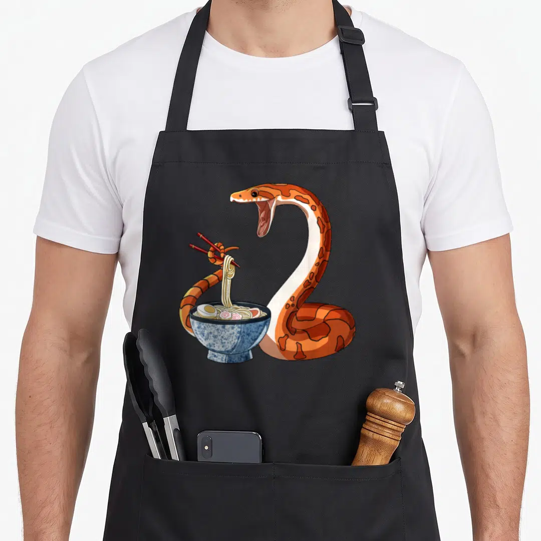 Japanese kawaii apron - Cartoon orange corn snake coiled around blue ramen bowl using tail to hold chopsticks — shown on a black cotton canvas apron against a white background