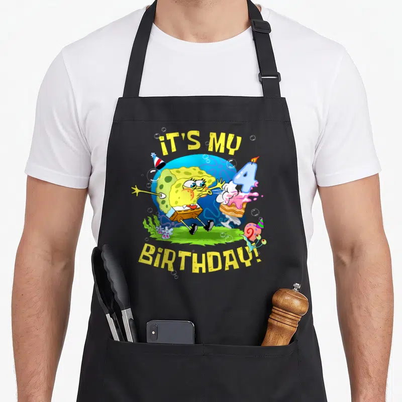 4th cake apron - Sponge character blowing number 4 candle on cake with IT'S MY BIRTHDAY text — shown on a black cotton canvas apron against a white background