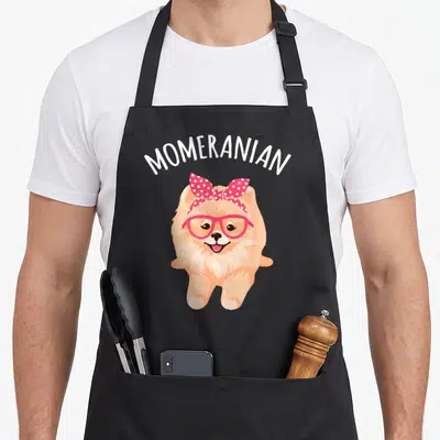 Mum pomeranian apron - Cartoon peach pomeranian wearing pink glasses and polka dot bow under white MOMERANIAN text — shown on a black cotton canvas apron against a white background