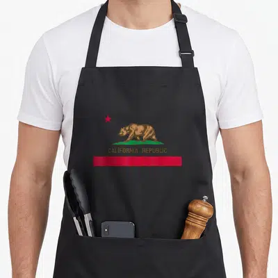 State california apron - Brown bear on green grass with red star and CALIFORNIA REPUBLIC text above a red stripe — shown on a black cotton canvas apron against a white background
