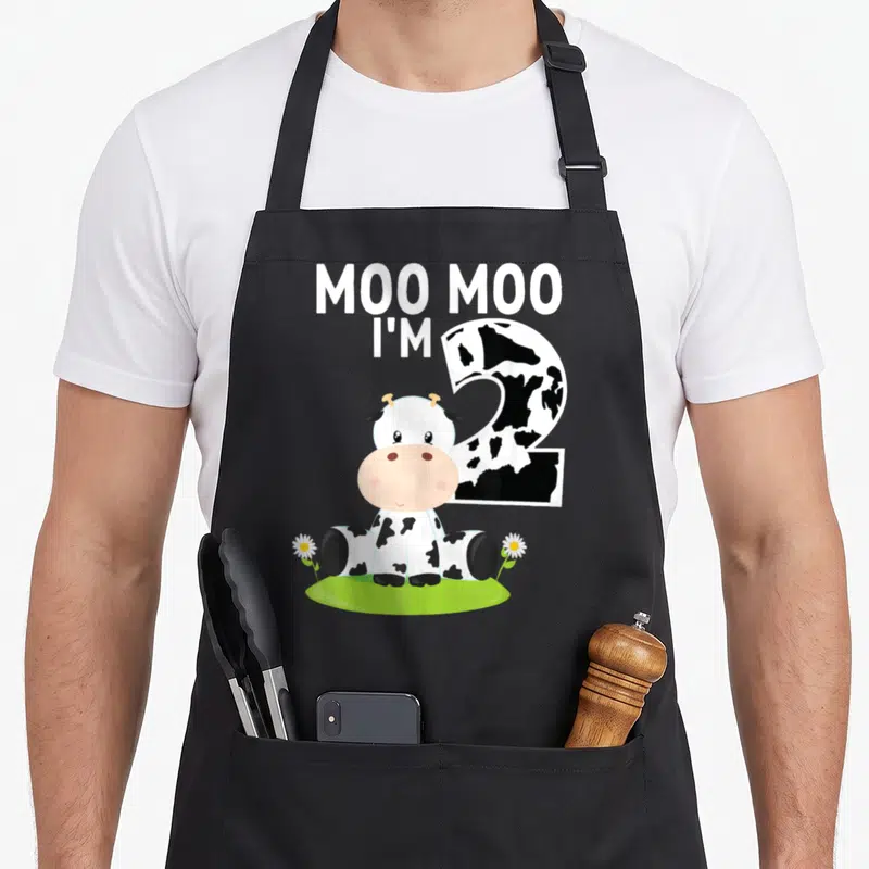 Cow 2nd apron - Moo Moo I'm Two text, spotted number two, sitting cow, daisies, green grass patch — shown on a black cotton canvas apron against a white background