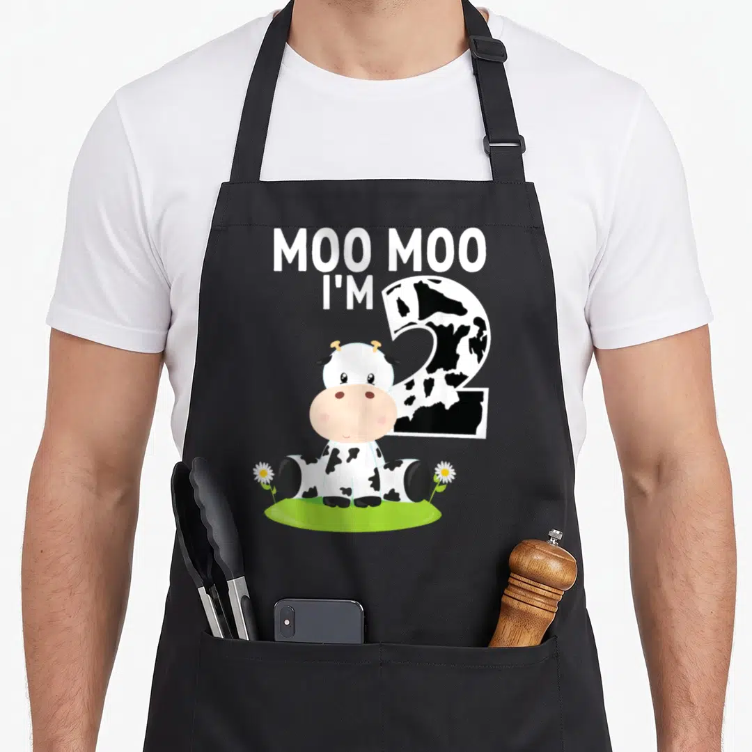 Cow 2nd apron - Moo Moo I'm Two text, spotted number two, sitting cow, daisies, green grass patch — shown on a black cotton canvas apron against a white background