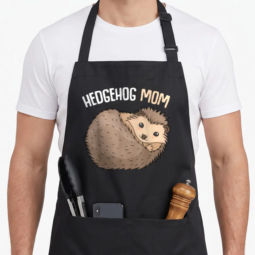 Cute funny hedgehog apron - Arched HEDGEHOG MOM text above a curled brown cartoon hedgehog — shown on a black cotton canvas apron against a white background