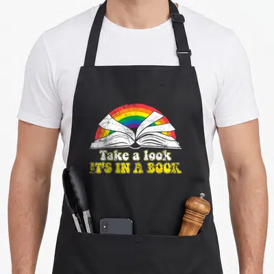 Reading rainbow apron - Open book under rainbow arch with Take a look IT'S IN A BOOK text in distressed colors — shown on a black cotton canvas apron against a white background