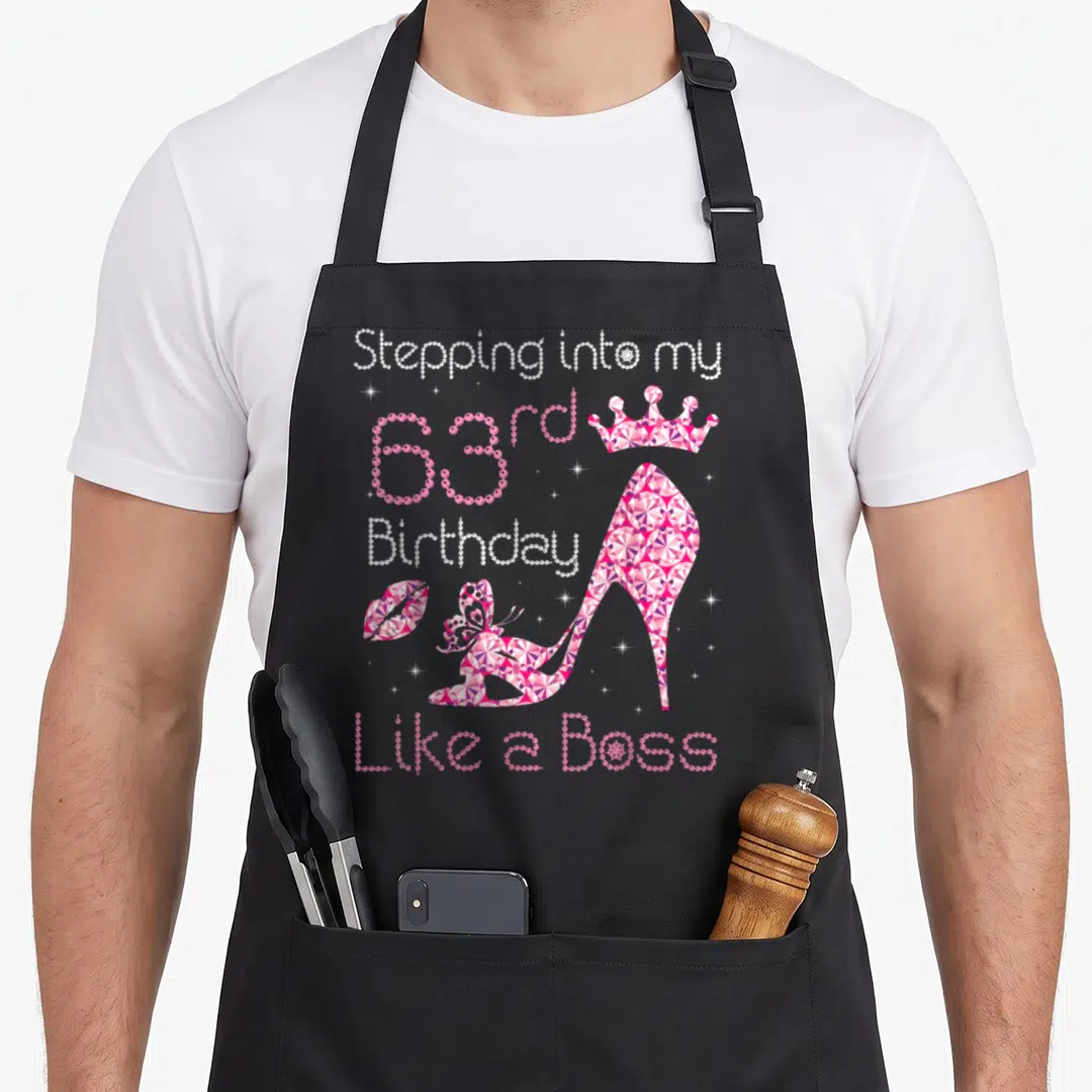 63rd bbq boss apron - Jeweled high heel shoe with crown, lips, butterfly, and 63rd birthday text in pink and purple — shown on a black cotton canvas apron against a white background