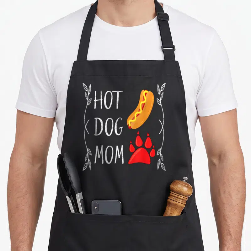 Eating hot dog apron - HOT DOG MOM text with cartoon hot dog and red paw print framed by leaves — shown on a black cotton canvas apron against a white background