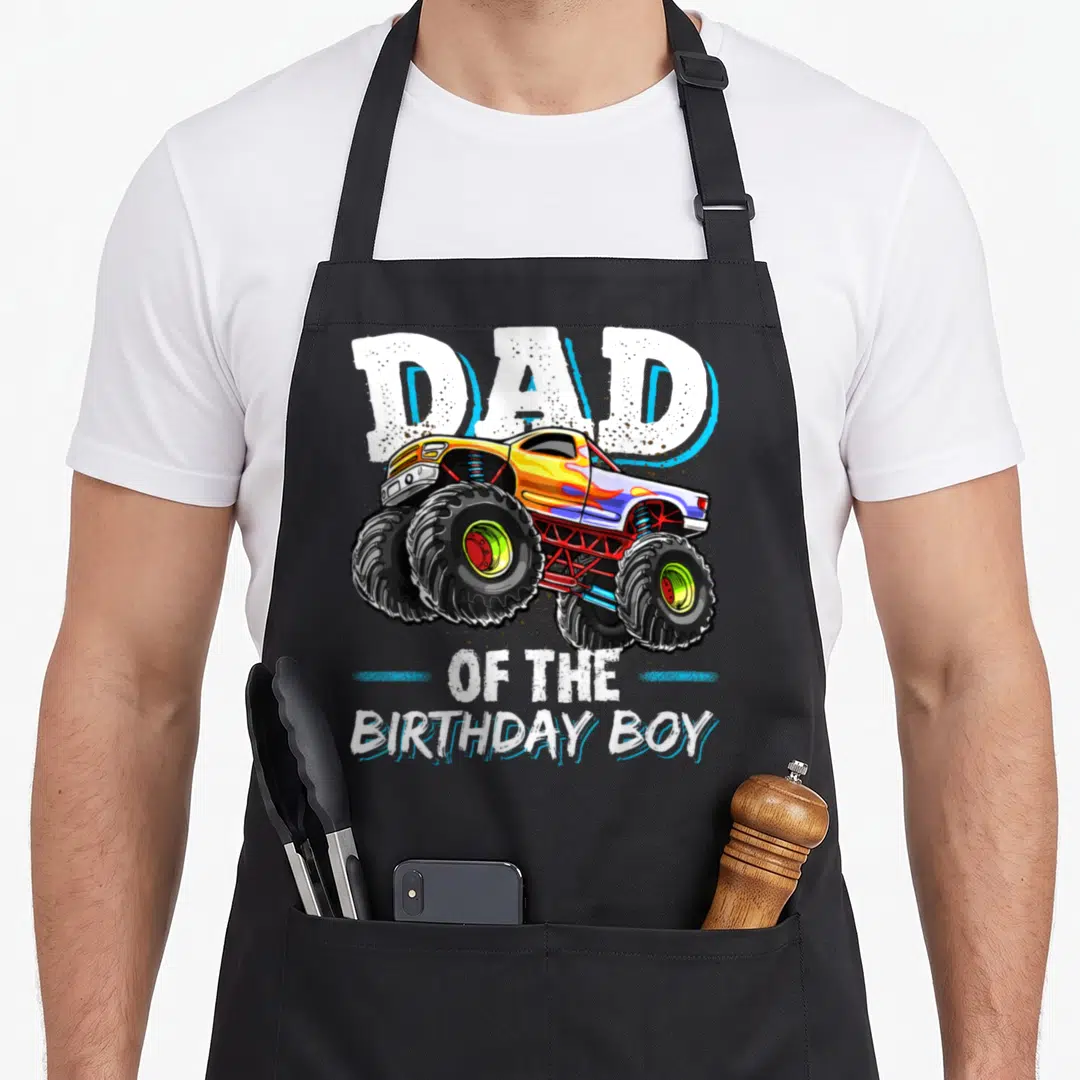 Birthday monster truck aprons - Cartoon monster truck with Dad of the Birthday Boy text in white and blue — shown on a black cotton canvas apron against a white background