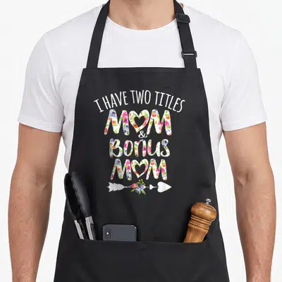 Title theme aprons - Stacked floral text reading I HAVE TWO TITLES MOM & BONUS MOM with heart arrow — shown on a black cotton canvas apron against a white background
