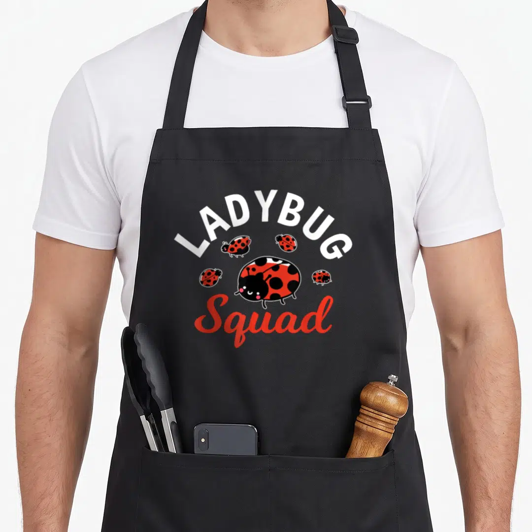 Vintage squad ladybug apron - Five red and black cartoon ladybugs clustered under white arching text and red script — shown on a black cotton canvas apron against a white background
