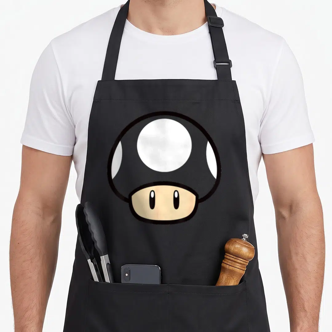 Funny face mushroom apron - Grey white and beige cartoon mushroom character with two eyes on a black bib — shown on a black cotton canvas apron against a white background