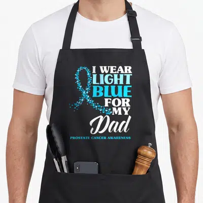 Light blue prostate cancer awareness apron - Ribbon formed from small blue hearts next to stacked white and blue text — shown on a black cotton canvas apron against a white background