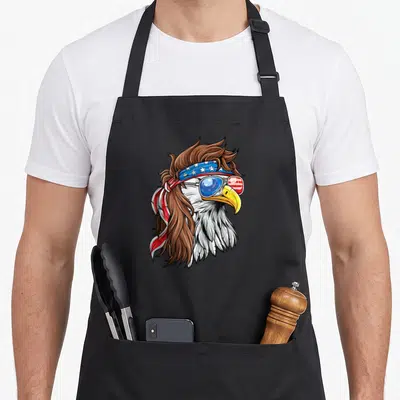 Eagle mullet 4th of july apron - Cartoon bald eagle with mullet, flag sunglasses, and star-spangled headband in red — shown on a black cotton canvas apron against a white background