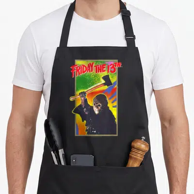 Vintage game aprons - FRIDAY THE 13TH text above masked figure holding axe on distressed colorful background — shown on a black cotton canvas apron against a white background