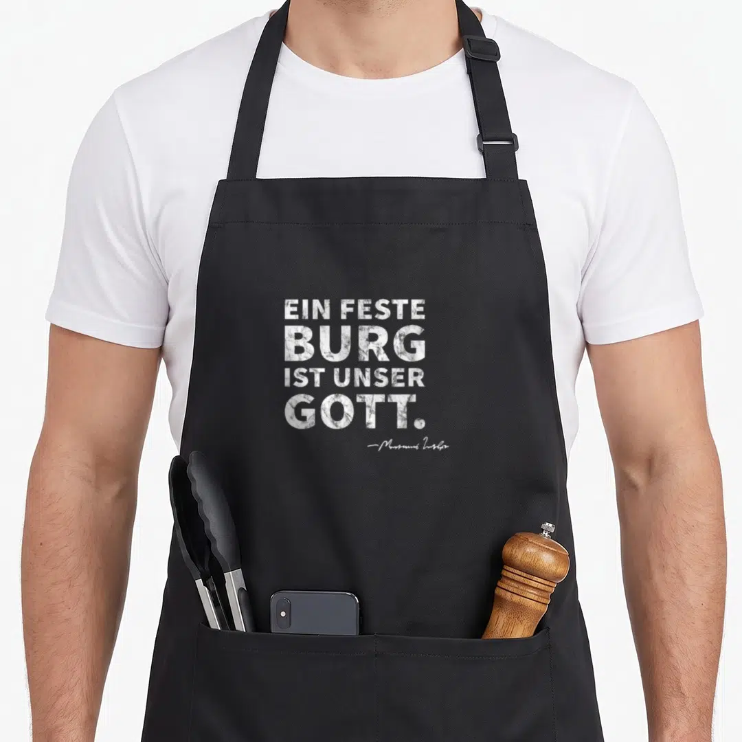 Mighty german apron - Distressed white German text EIN FESTE BURG IST UNSER GOTT. with Martin Luther signature — shown on a black cotton canvas apron against a white background