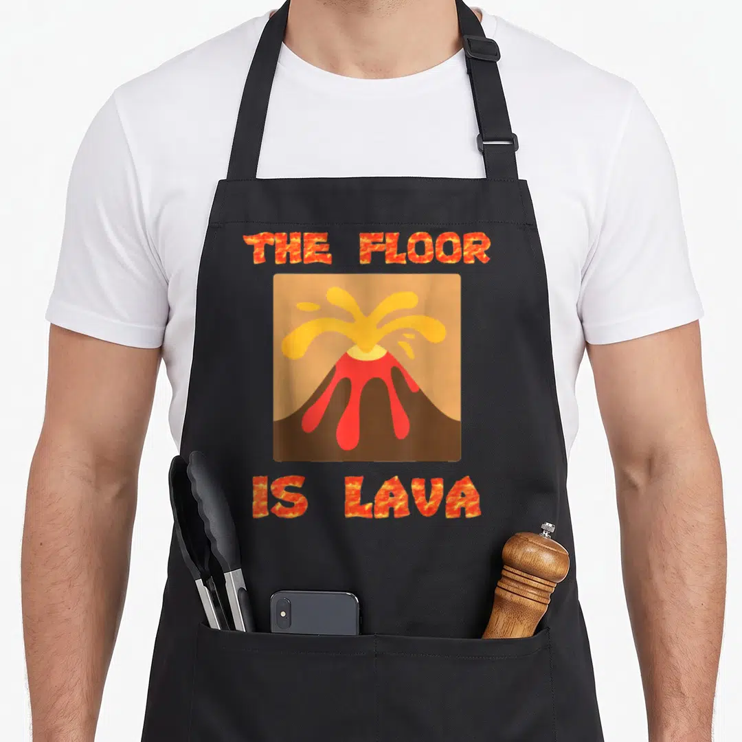 Floor geek apron - Cartoon volcano erupting red and orange lava between the words THE FLOOR IS LAVA in textured font — shown on a black cotton canvas apron against a white background