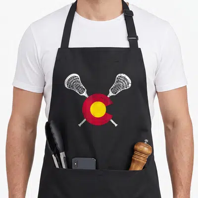 Lacrosse colorado apron - Crossed white lacrosse sticks over distressed red circle and yellow center on black apron — shown on a black cotton canvas apron against a white background