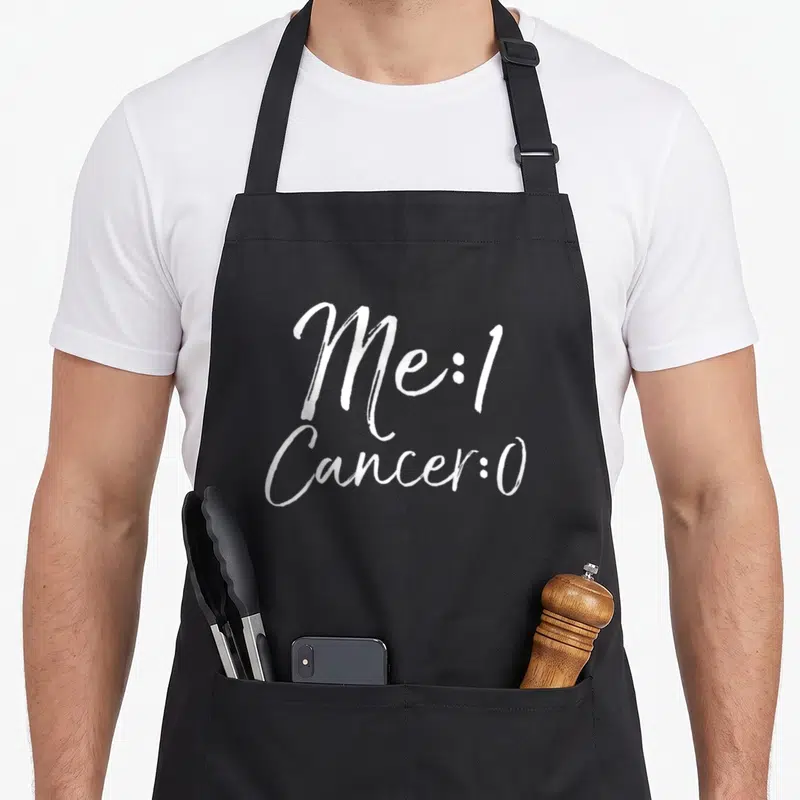 Cancer remission apron - White script text Me:1 Cancer:0 stacked vertically — shown on a black cotton canvas apron against a white background