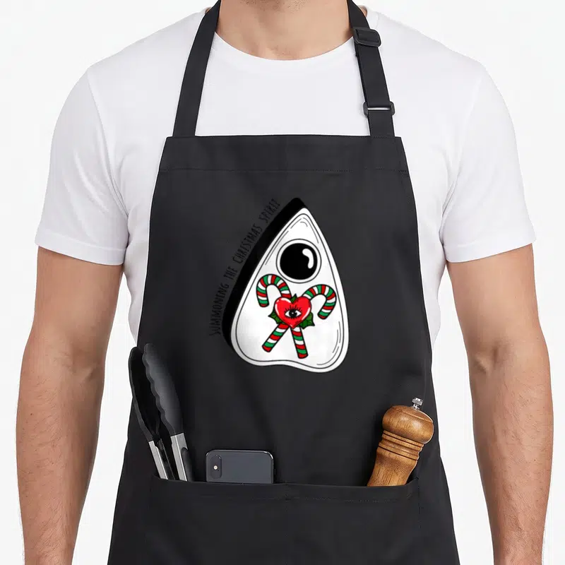 Ugly retro christmas aprons - Ouija planchette with SUMMONING THE CHRISTMAS SPIRIT text and candy cane eye art — shown on a black cotton canvas apron against a white background