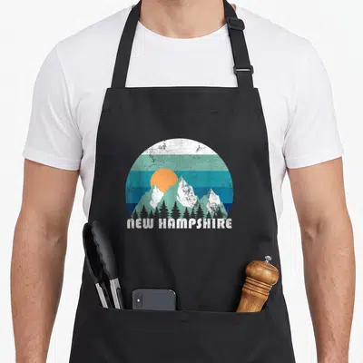 State hampshire girl apron - Retro New Hampshire mountain landscape with orange sun, striped sky, and trees — shown on a black cotton canvas apron against a white background