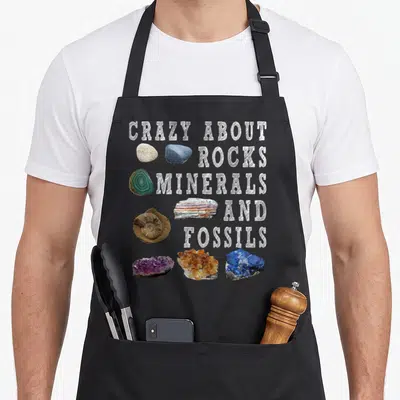 Mineral teacher apron - Stacked white text next to purple, blue, orange, and grey rock specimens on black background — shown on a black cotton canvas apron against a white background