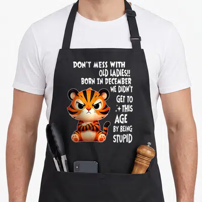 Mess tiger lady apron - Cartoon tiger with crossed arms and white text warning not to mess with old ladies born — shown on a black cotton canvas apron against a white background