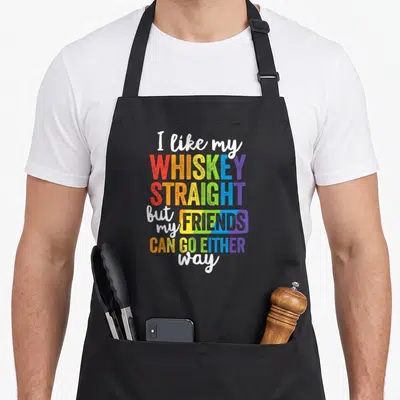 Whiskey gay pride apron - Rainbow text reading I like my WHISKEY STRAIGHT but my FRIENDS CAN GO EITHER way on black — shown on a black cotton canvas apron against a white background