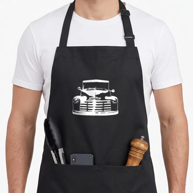 Hot truck apron - white line-art front view of vintage truck with CHEVROLET text on grille — shown on a black cotton canvas apron against a white background