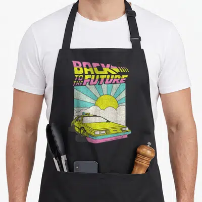 Delorean cartoon apron - Back to the Future text above yellow car with pink blue sun rays — shown on a black cotton canvas apron against a white background