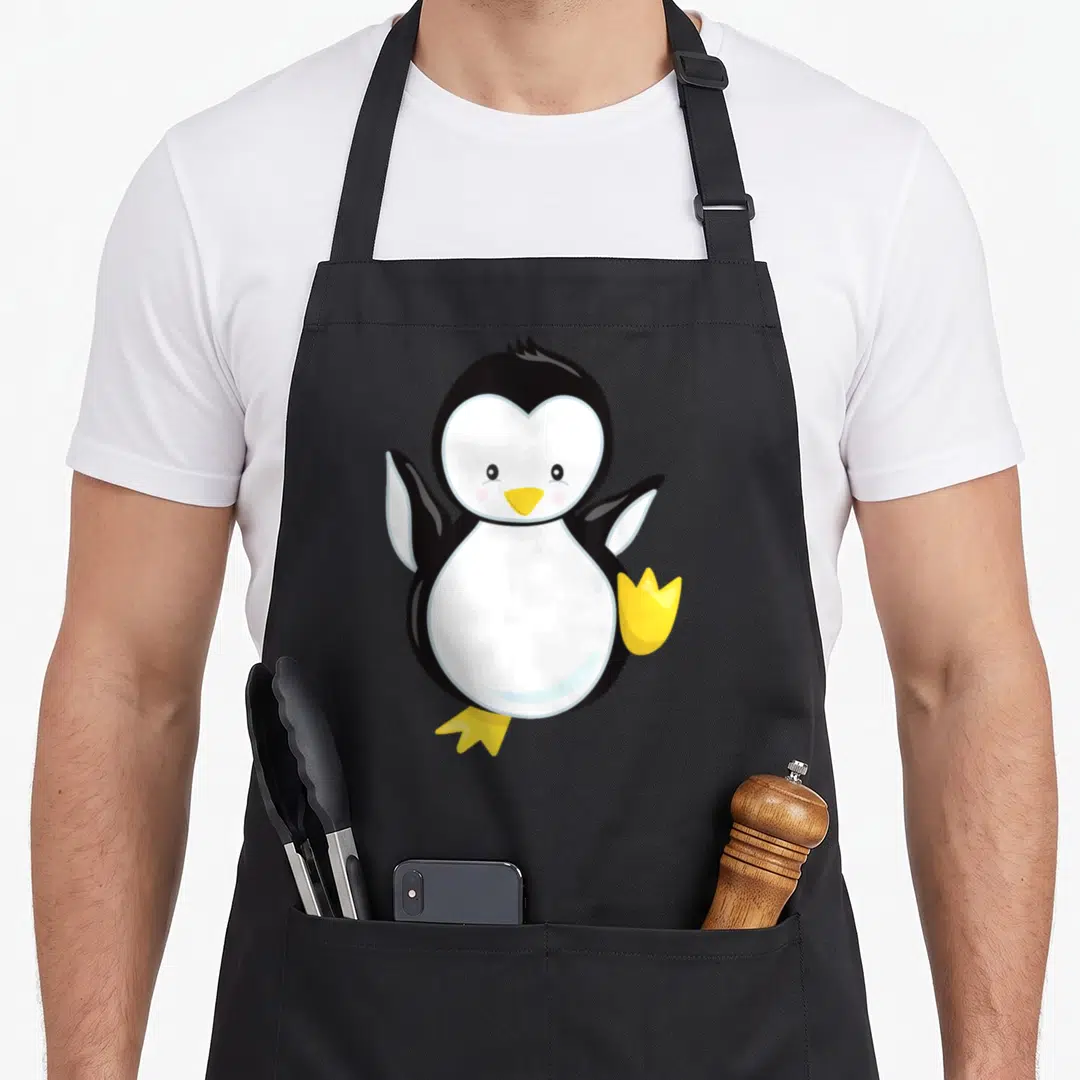 Modern trendy apron - Cartoon penguin with raised wings, yellow beak and feet, black and white body, and pink cheeks — shown on a black cotton canvas apron against a white background