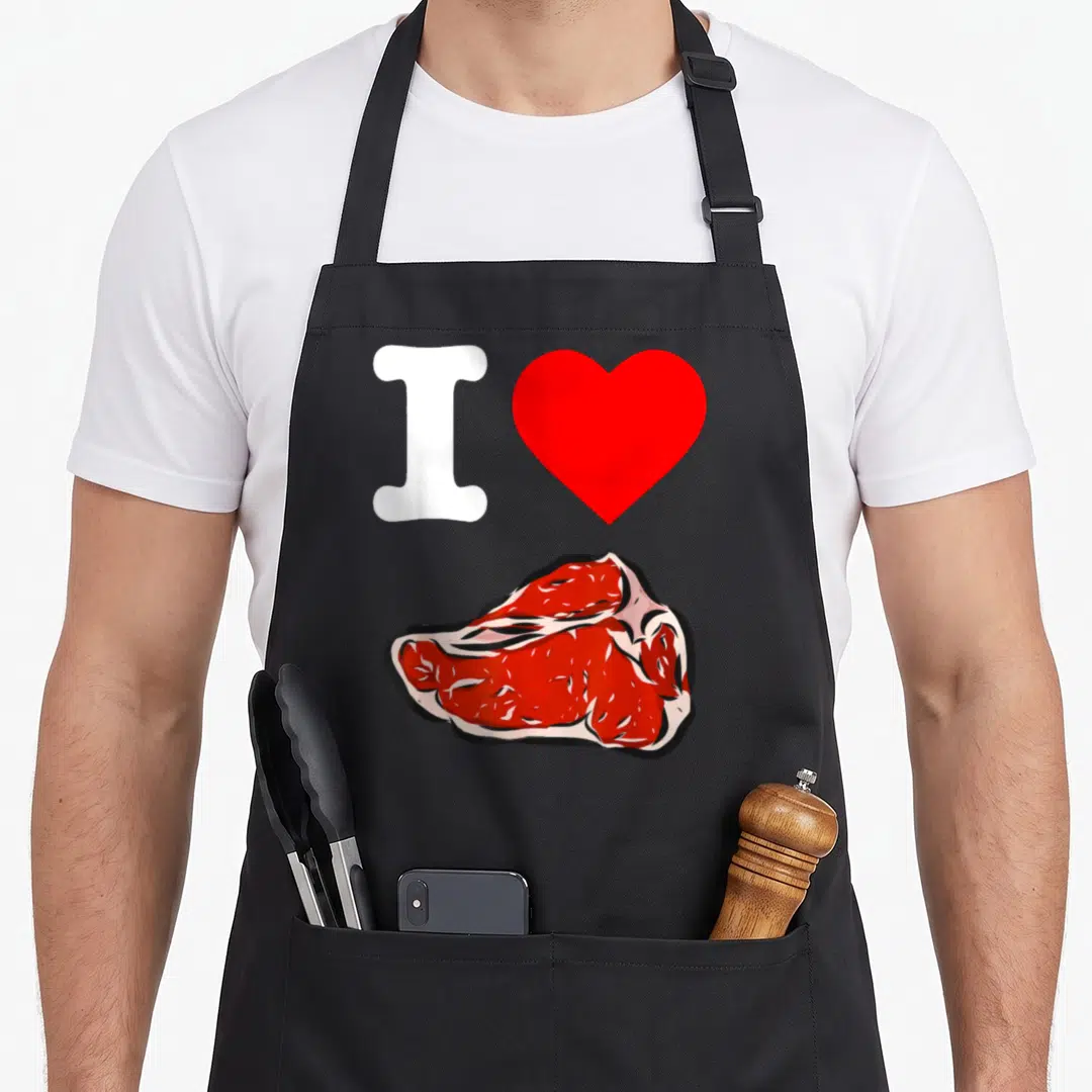 Meat manly aprons - White letter I and red heart above illustrated raw red meat block — shown on a black cotton canvas apron against a white background