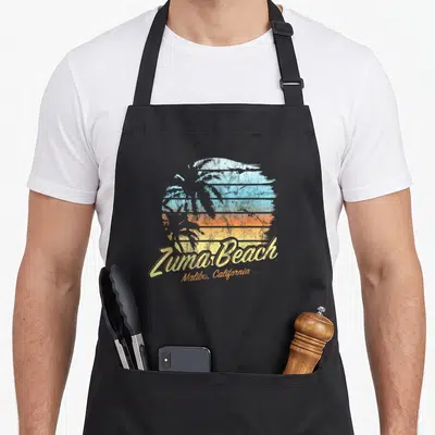 Malibu zuma beach apron - Retro circular sunset with palm tree silhouette and Zuma Beach Malibu California text in blue — shown on a black cotton canvas apron against a white background