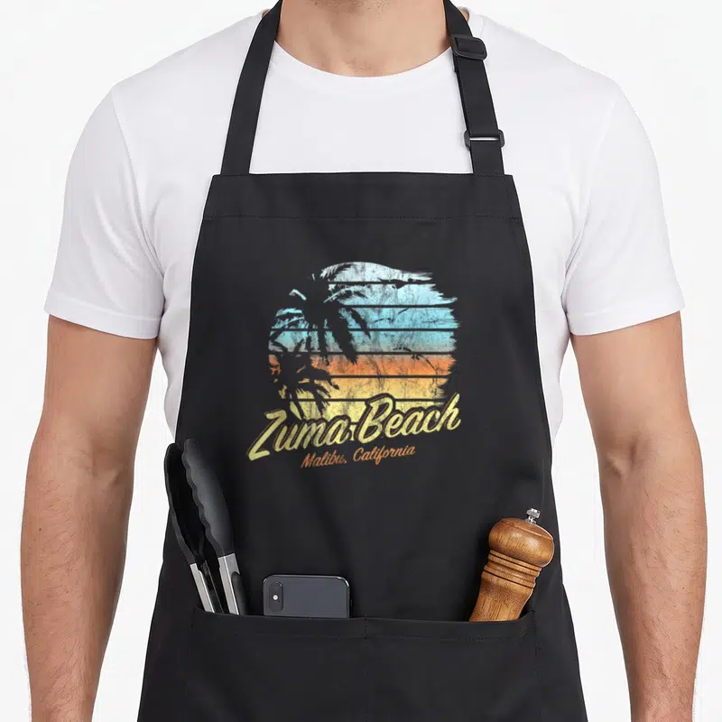 Malibu zuma beach apron - Retro circular sunset with palm tree silhouette and Zuma Beach Malibu California text in blue — shown on a black cotton canvas apron against a white background